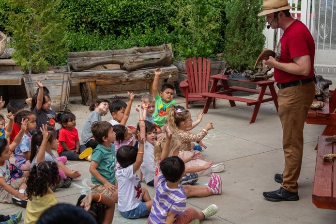 Children raising their hands at an outdoor dinosaur assembly with a naturalist